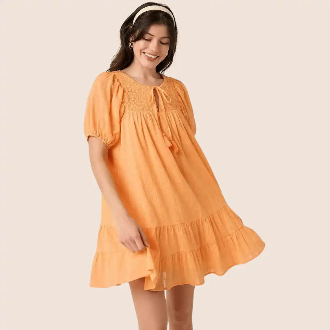 A woman in a short, tiered, ruffle-hemmed orange smock dress with puffed sleeves and a tie-front neckline.